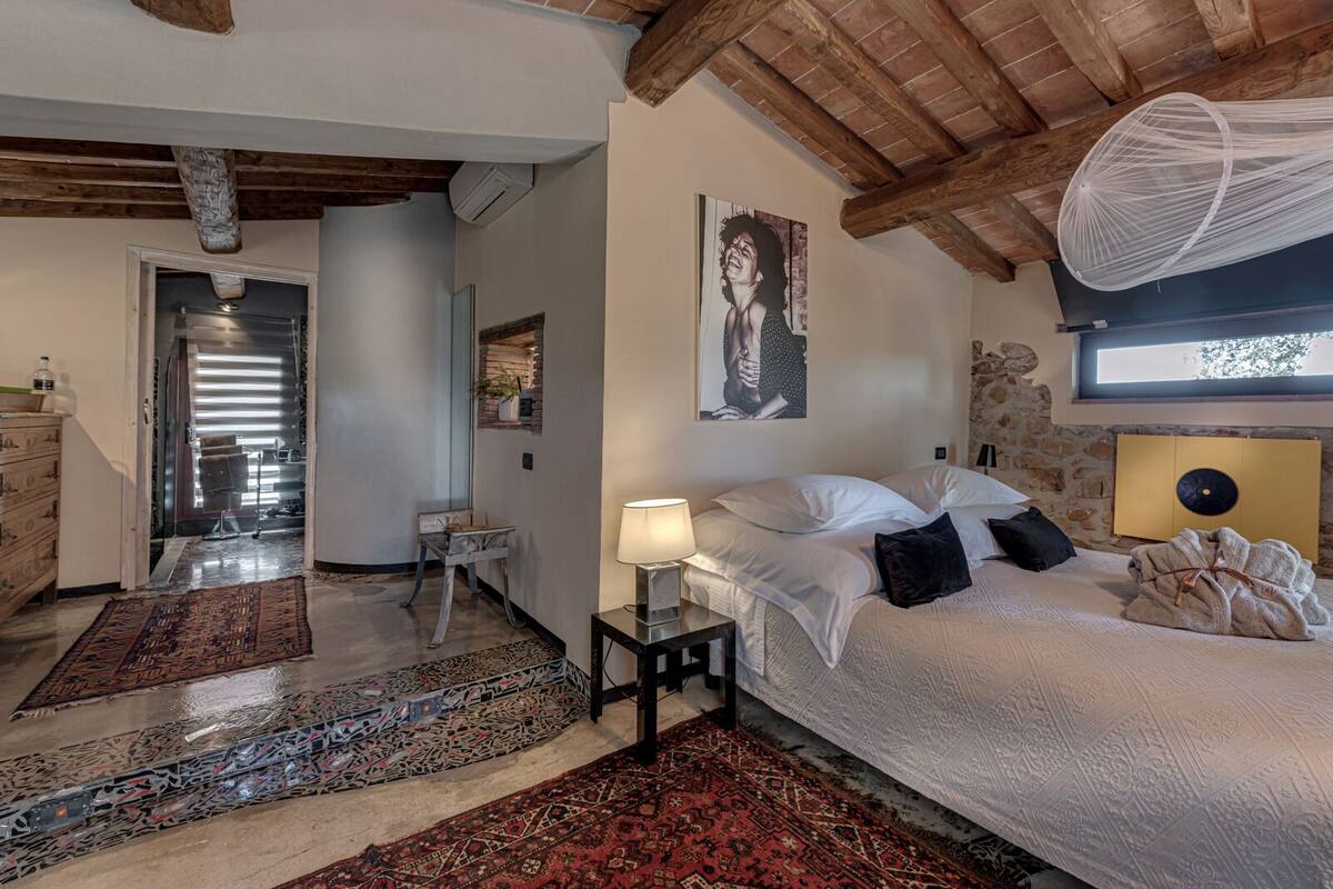 A bedroom with a large bed, a bedside table with a lamp, a hanging light fixture, a stone wall, and a window with a view.