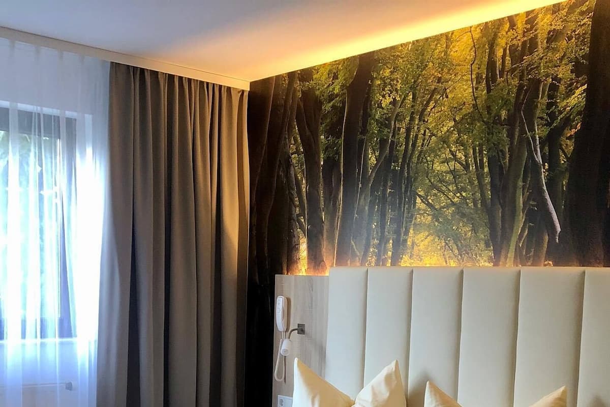 Une chambre d’hôtel avec un grand lit, deux oreillers, une tête de lit dotée d’un téléphone et une fresque représentant une forêt sur le mur.