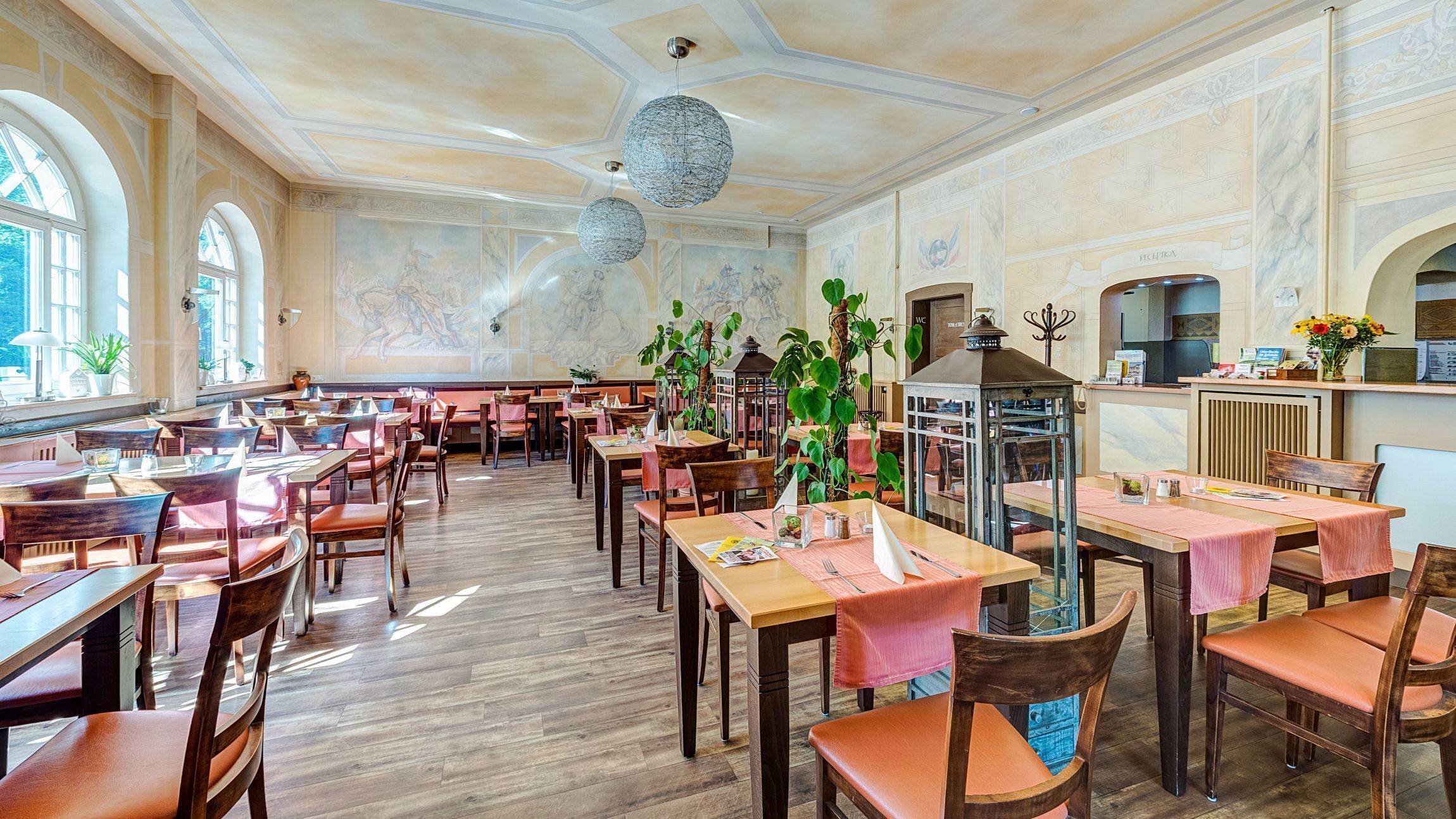 A spacious restaurant with wooden tables and chairs, large windows, and decorative ceiling elements.