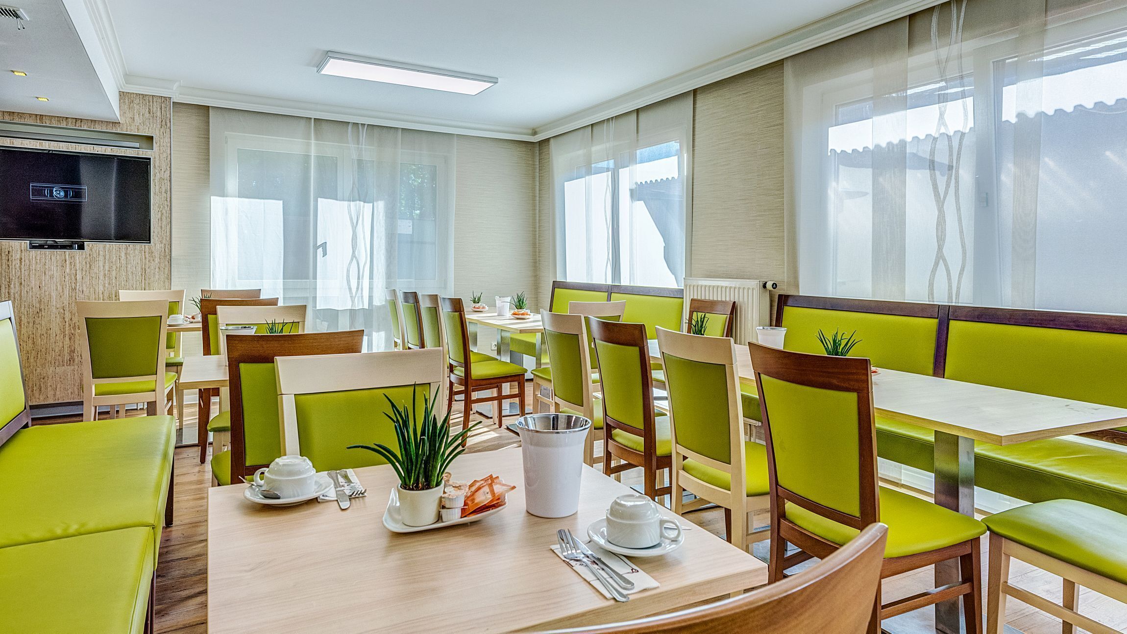 A hotel dining area with tables and chairs, a television, and large windows.