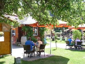 An outdoor seating area with wicker chairs and tables, surrounded by greenery and a building with a sloped roof. - Hotel & Restaurant Zur Kanone (Tautenhain)