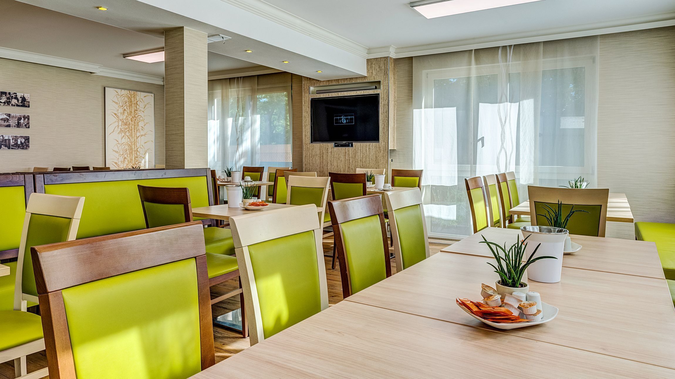 A modern dining area with green upholstered chairs, wooden tables, and a flat-screen TV mounted on the wall.