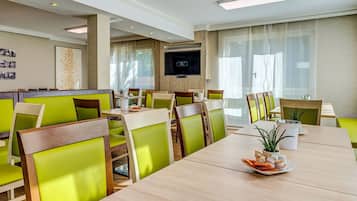 A modern dining area with green upholstered chairs, wooden tables, and a flat-screen TV mounted on the wall.