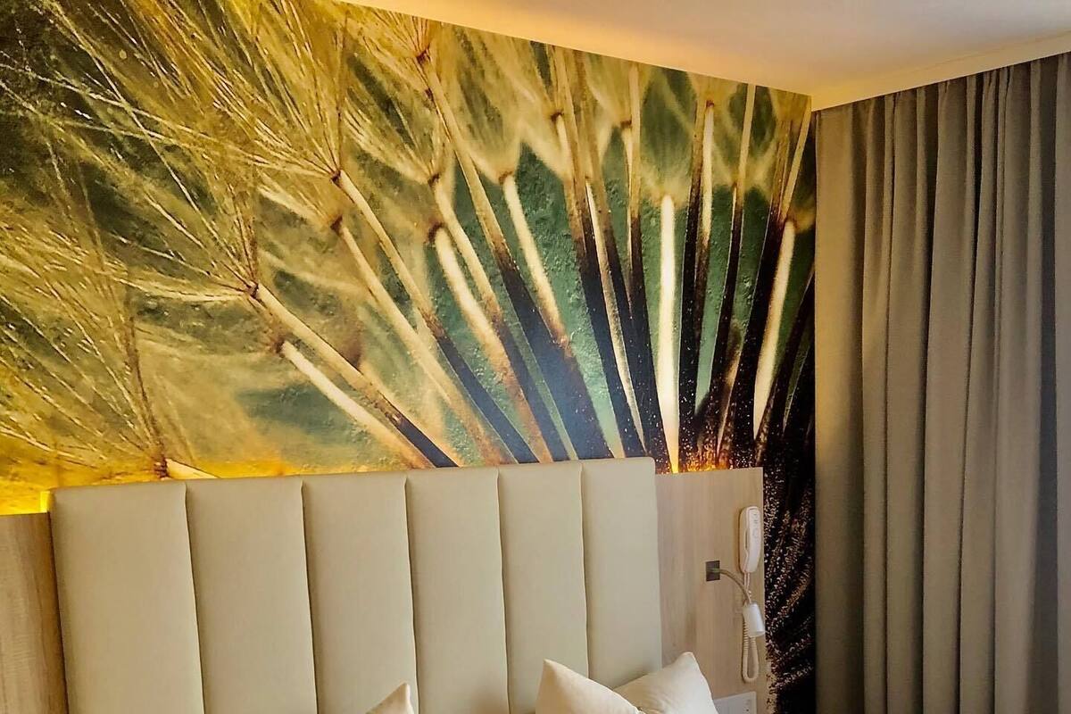 Une chambre d’hôtel avec un grand lit, deux oreillers, une table de chevet, une lampe et un mur orné d’un motif de plumes de paon.