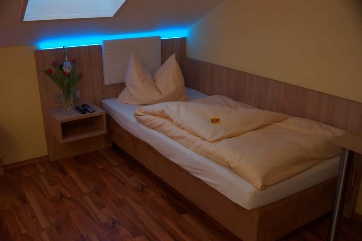 A bedroom with a wooden bed, a nightstand, a small table with a vase of flowers, and a wall-mounted shelf.