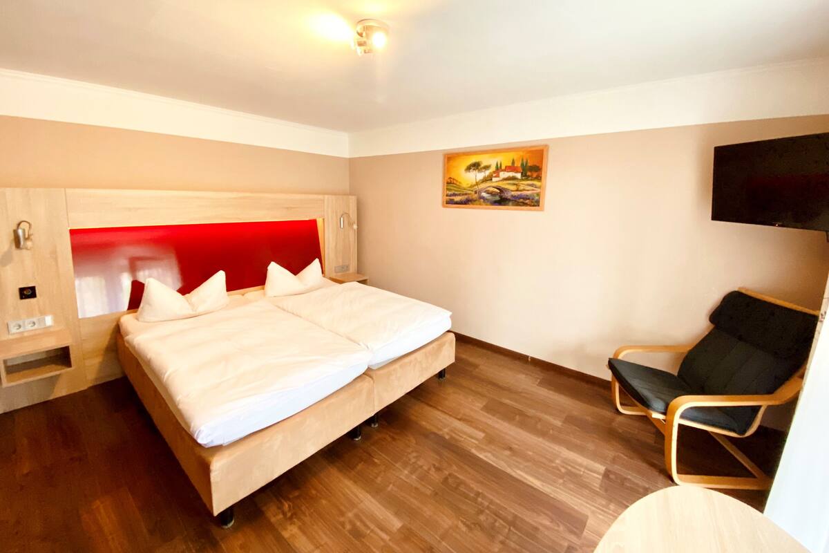 A modern hotel room with a large bed, a wooden headboard, a bedside table, a chair, and a painting on the wall.
