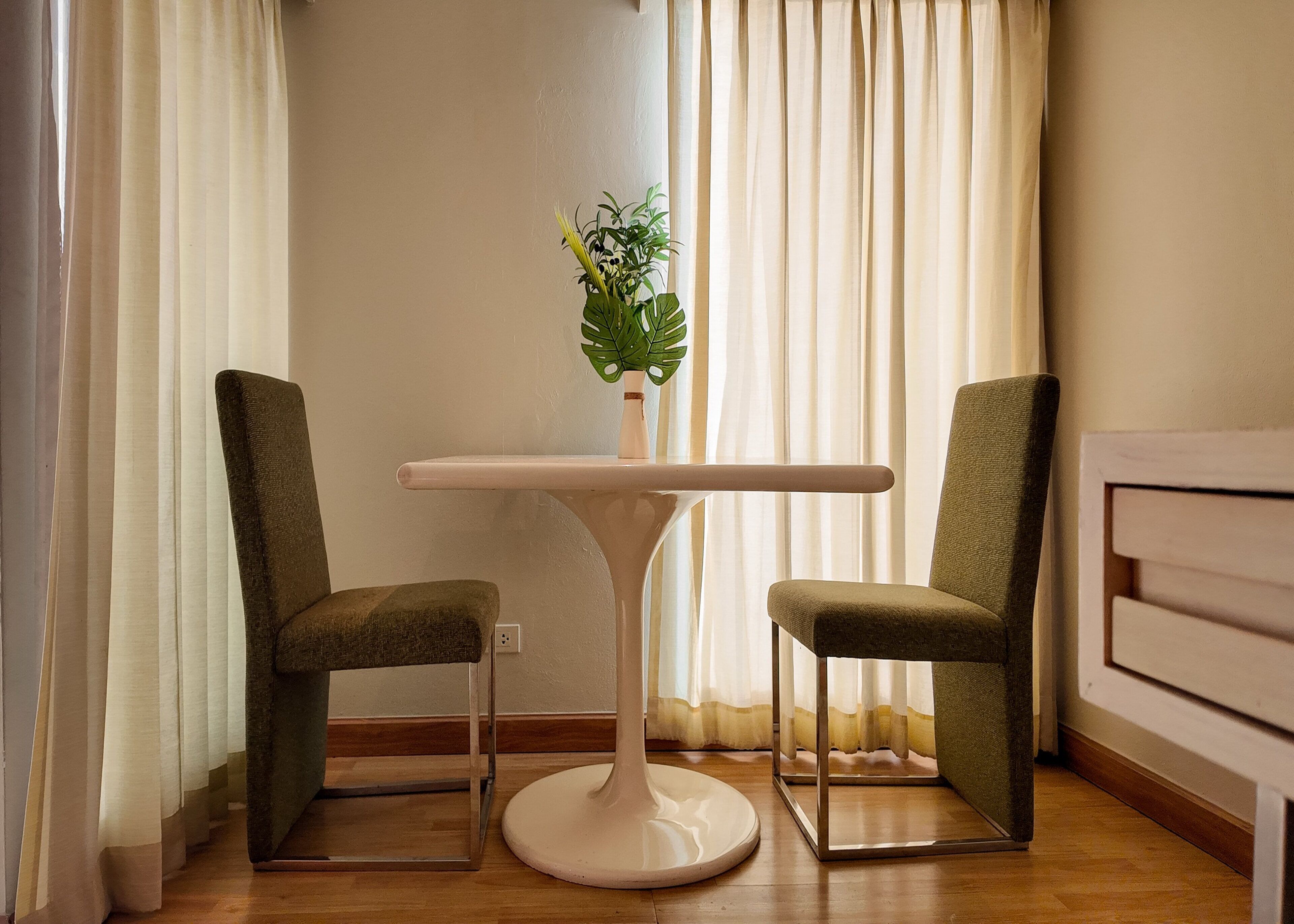 A room with a white round table, two green upholstered chairs, a potted plant on the table, and sheer curtains.