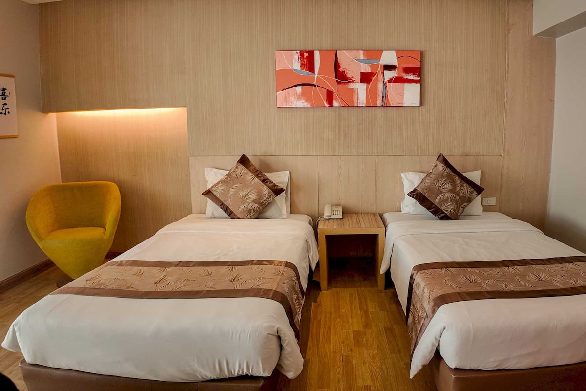 A hotel room with two beds, a bedside table with a phone, a lamp, and a yellow armchair.