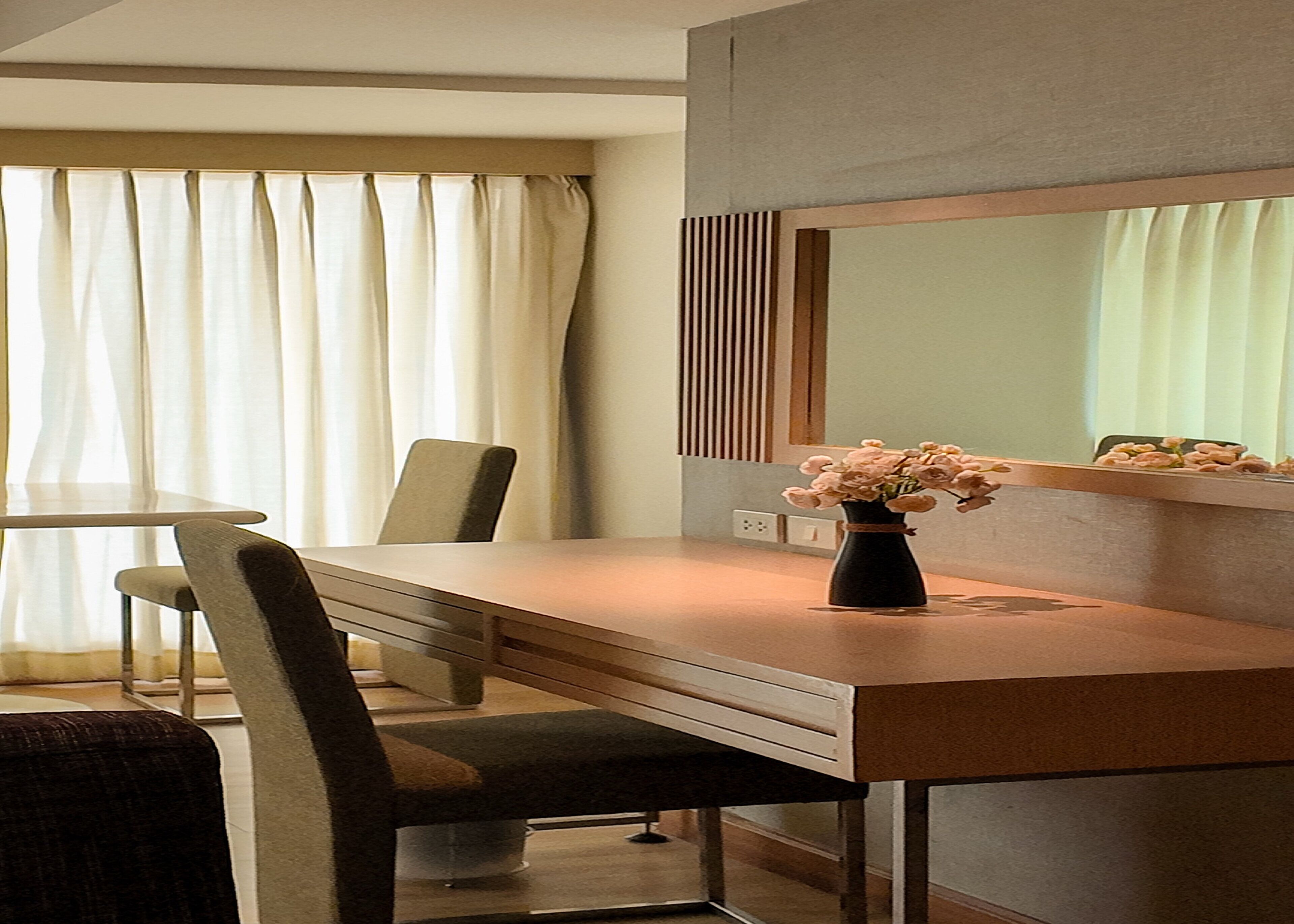 A hotel room with a wooden desk, a chair, a vase with flowers, a mirror, and a window with curtains.