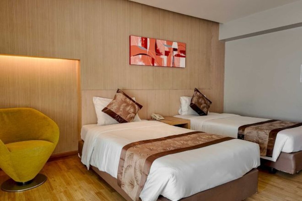 A hotel room with two beds, a yellow armchair, a bedside table, and a wall-mounted artwork.