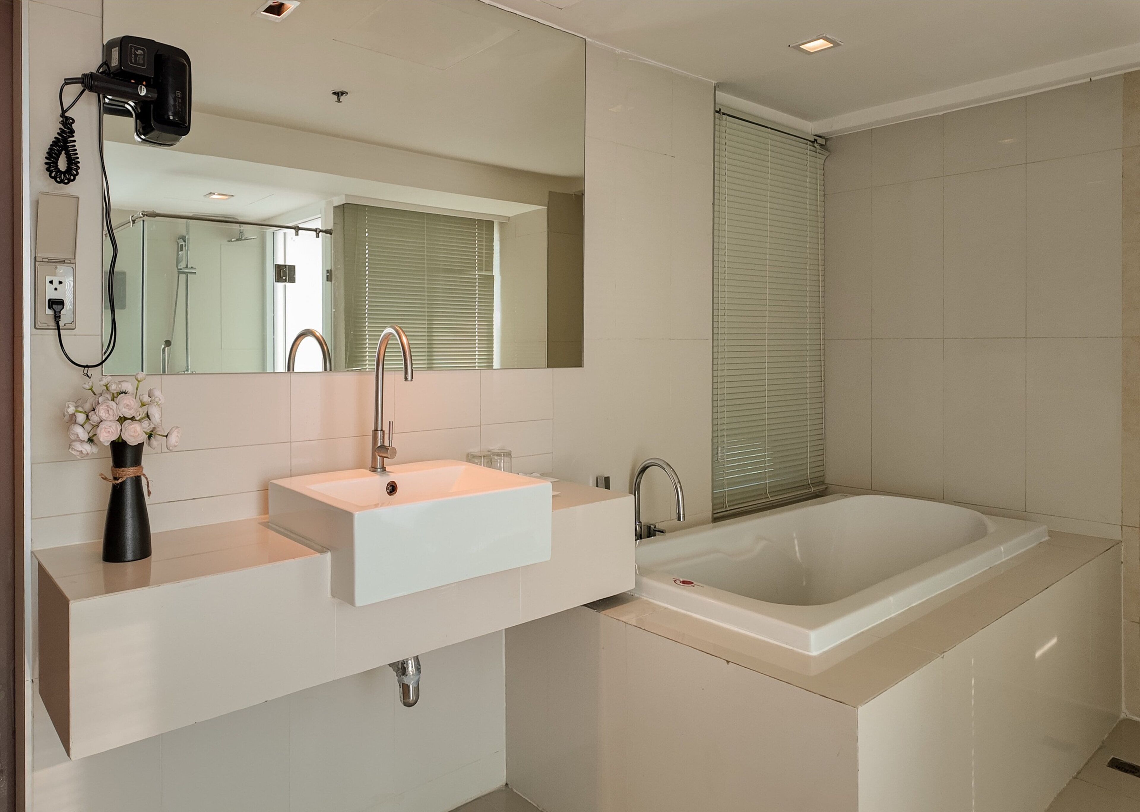 A modern bathroom with a white sink, a large mirror, a hairdryer, and a bathtub.
