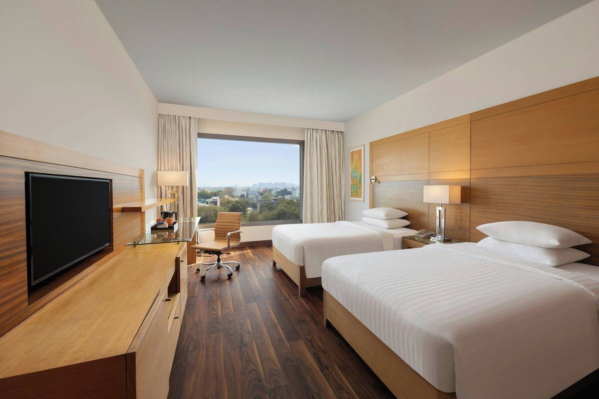 Ein Hotelzimmer mit großem Fernseher, zwei Betten, einem Schreibtisch mit Stuhl, einer Lampe und Blick auf die Stadt.