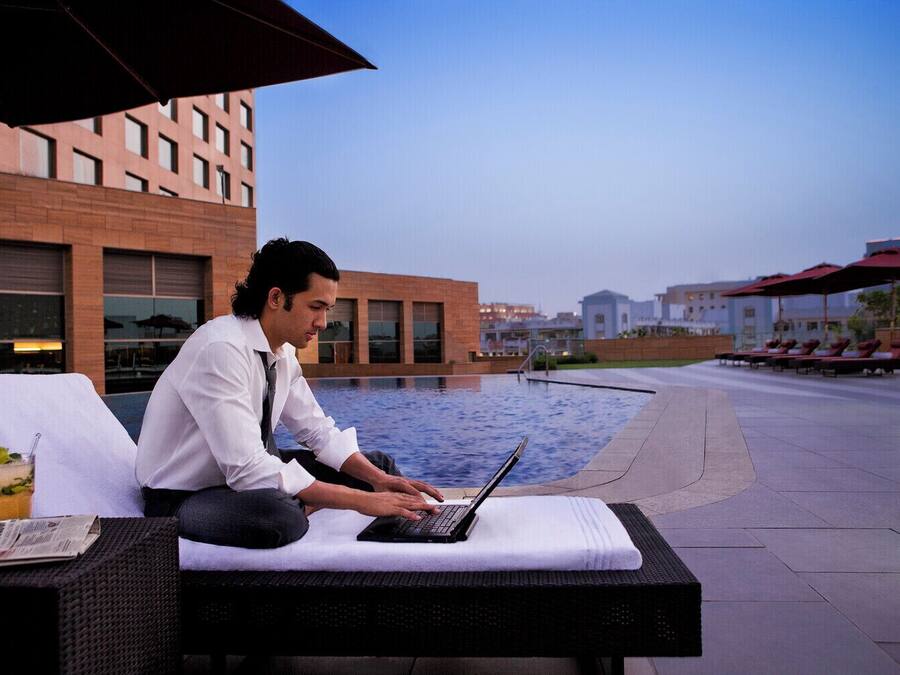 A man sitting on a lounge chair by a pool with a laptop, a drink, and a newspaper on the table.
