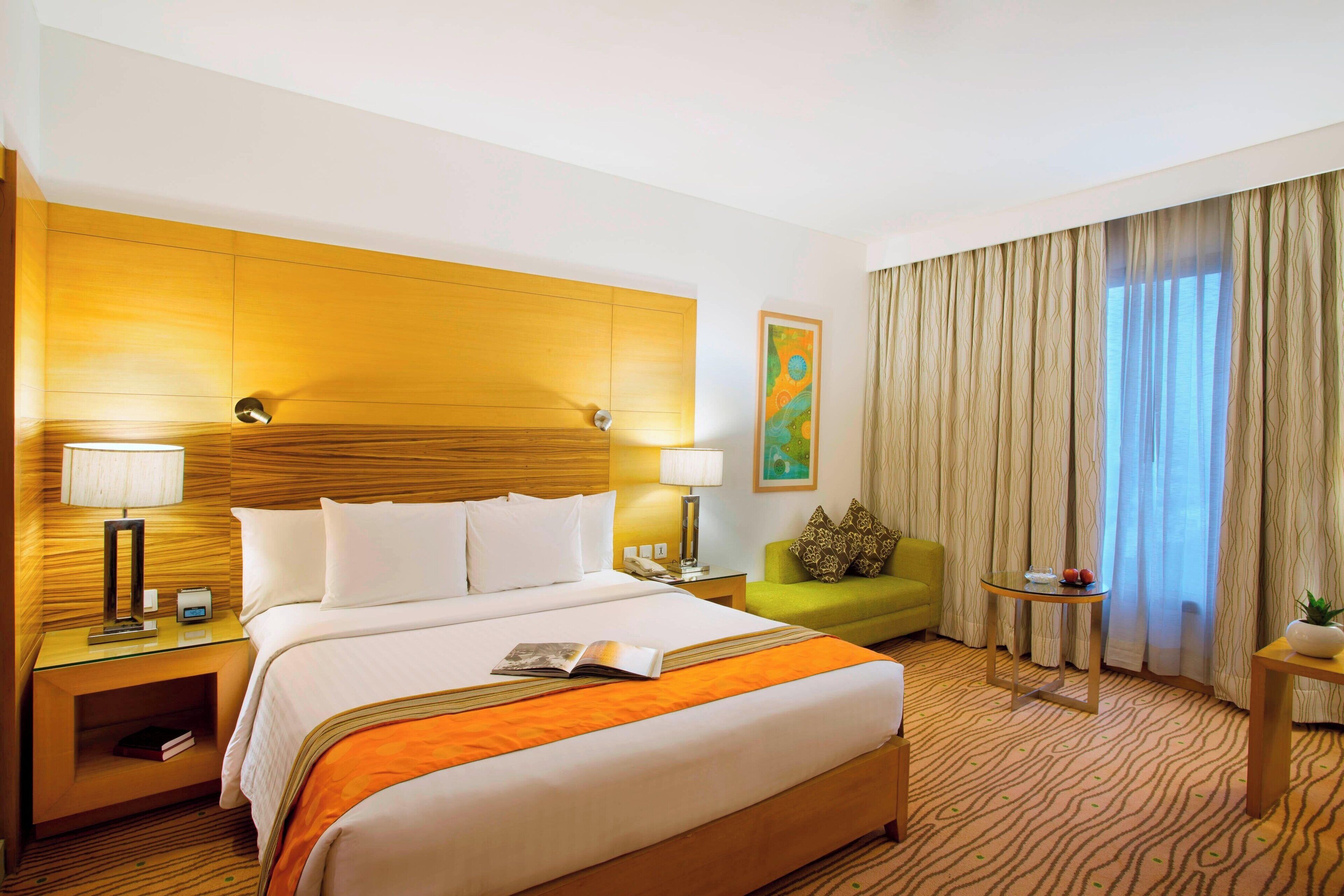 A hotel room with a large bed, two bedside tables with lamps, a green armchair, a small table with a plant, and a window with curtains.