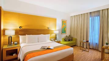 A hotel room with a large bed, two bedside tables with lamps, a green armchair, a small table with a plant, and a window with curtains.