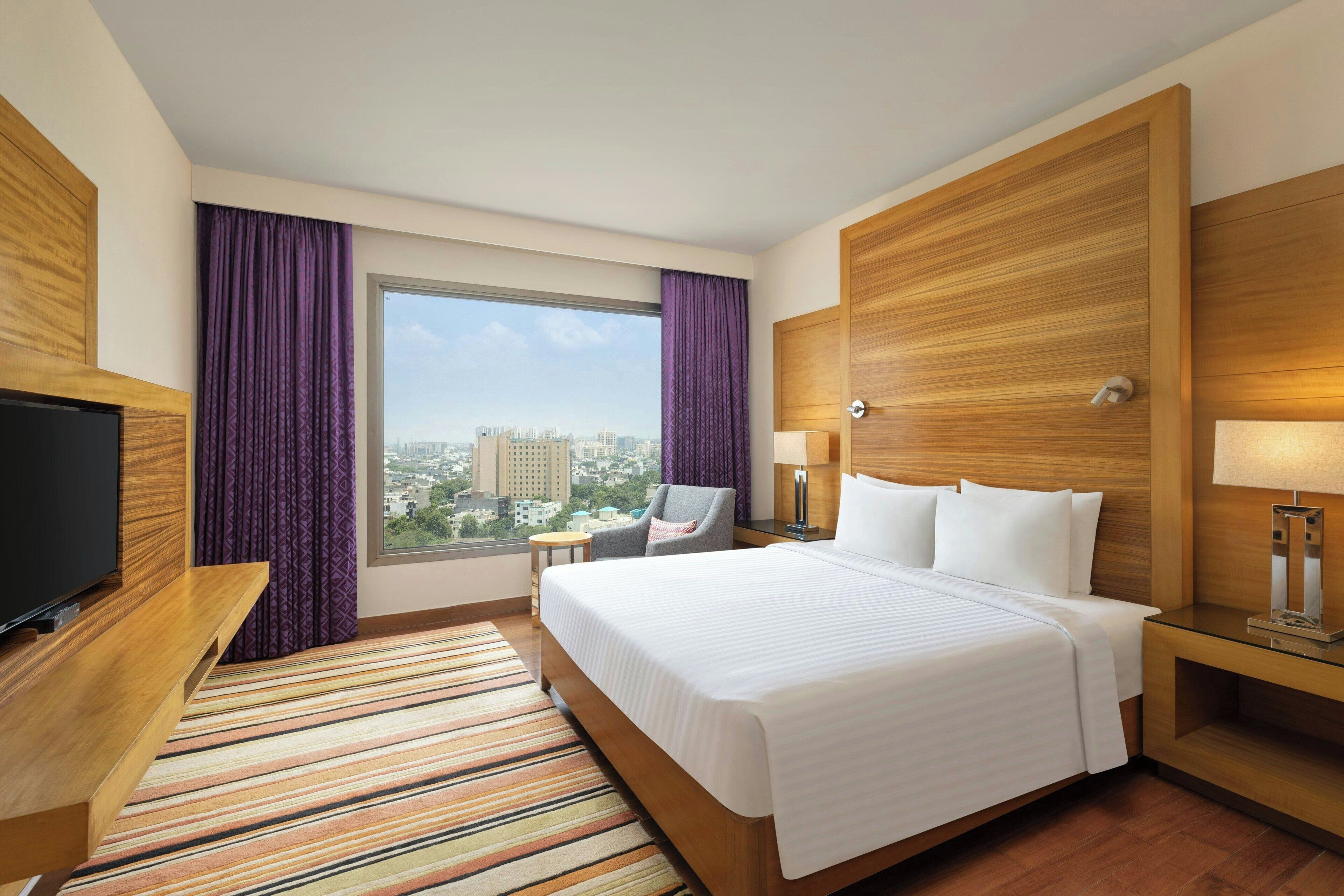 A hotel room with a large bed, a TV, a desk, a chair, a window with a city view, and a wooden headboard.