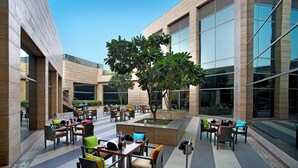 An outdoor dining area with multiple tables and chairs, surrounded by a modern building with large windows and a central tree. - Courtyard by Marriott Gurugram Downtown (Gurugram)