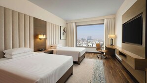 A hotel room with two beds, a large TV, a desk with a chair, and a view of the city. - Courtyard by Marriott Gurugram Downtown (Gurugram)