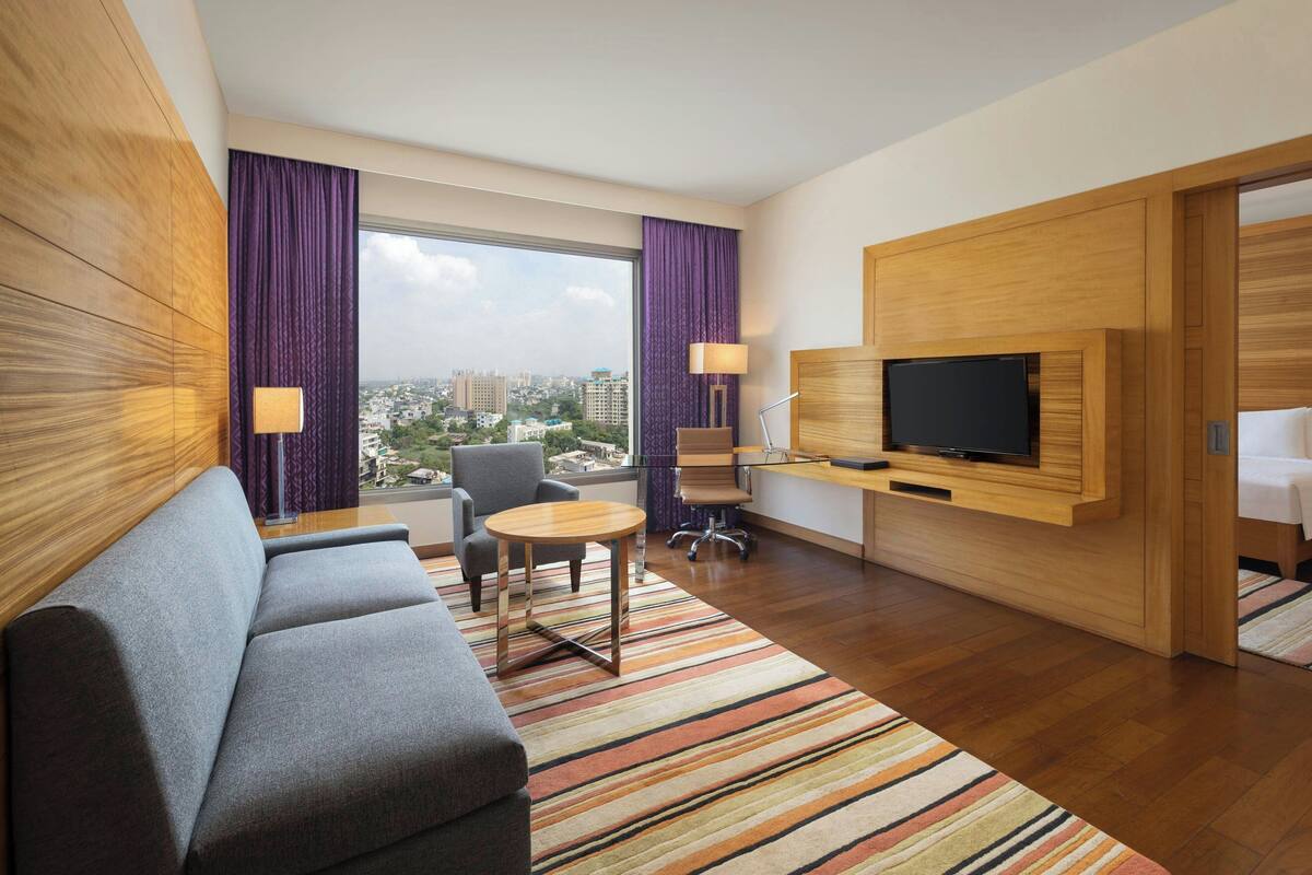 Ein modernes Hotelzimmer mit Sofa, kleinem runden Tisch, Wandfernseher und großem Fenster mit Vorhängen.