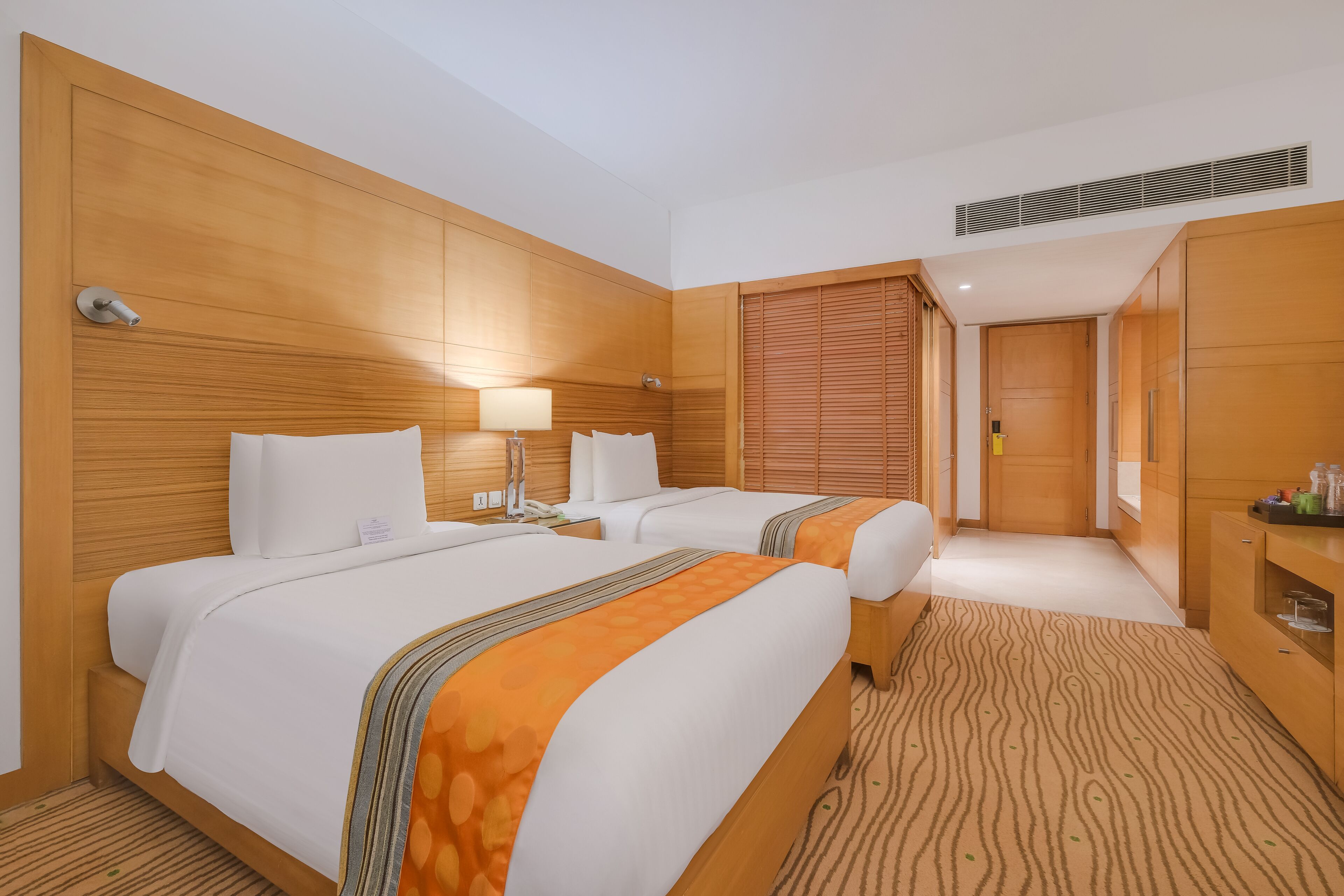 Quarto de hotel com duas camas, cabeceiras de madeira, mesa de cabeceira com luminária, guarda-roupa e carpete estampado.