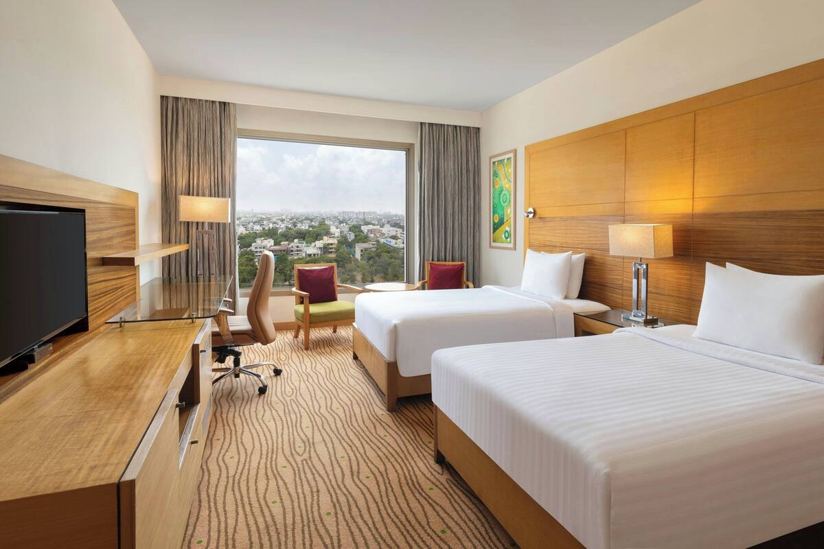 Ein Hotelzimmer mit zwei Betten, einem Schreibtisch mit Fernseher, einem Sessel und einem großen Fenster mit Blick auf die Stadt.