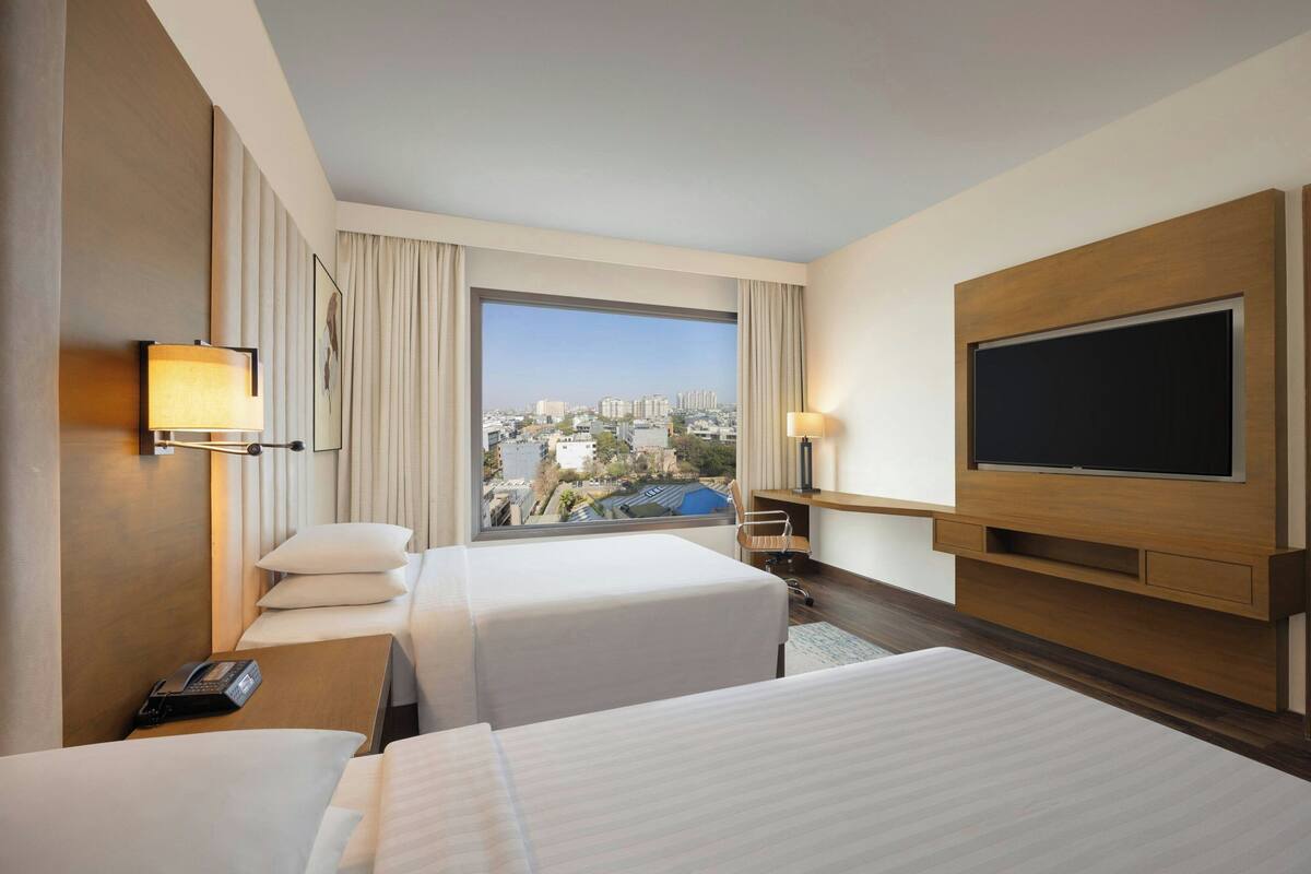Ein Hotelzimmer mit zwei Betten, einem großen Fenster mit Blick auf die Stadt, einem Flachbildfernseher und einem Schreibtisch mit Lampe.
