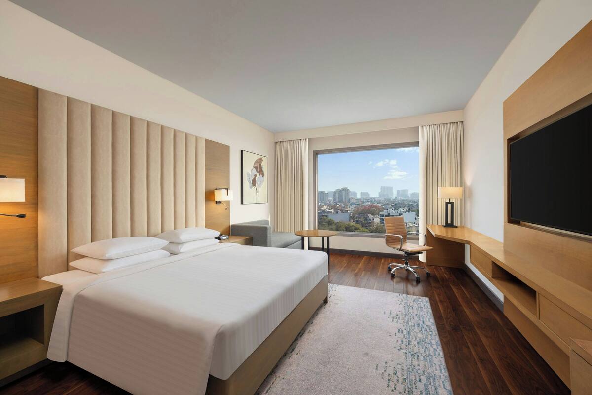Ein Hotelzimmer mit einem großen Bett, einem Schreibtisch, einem Sessel, einem Fernseher und einem Fenster mit Blick auf die Stadt.