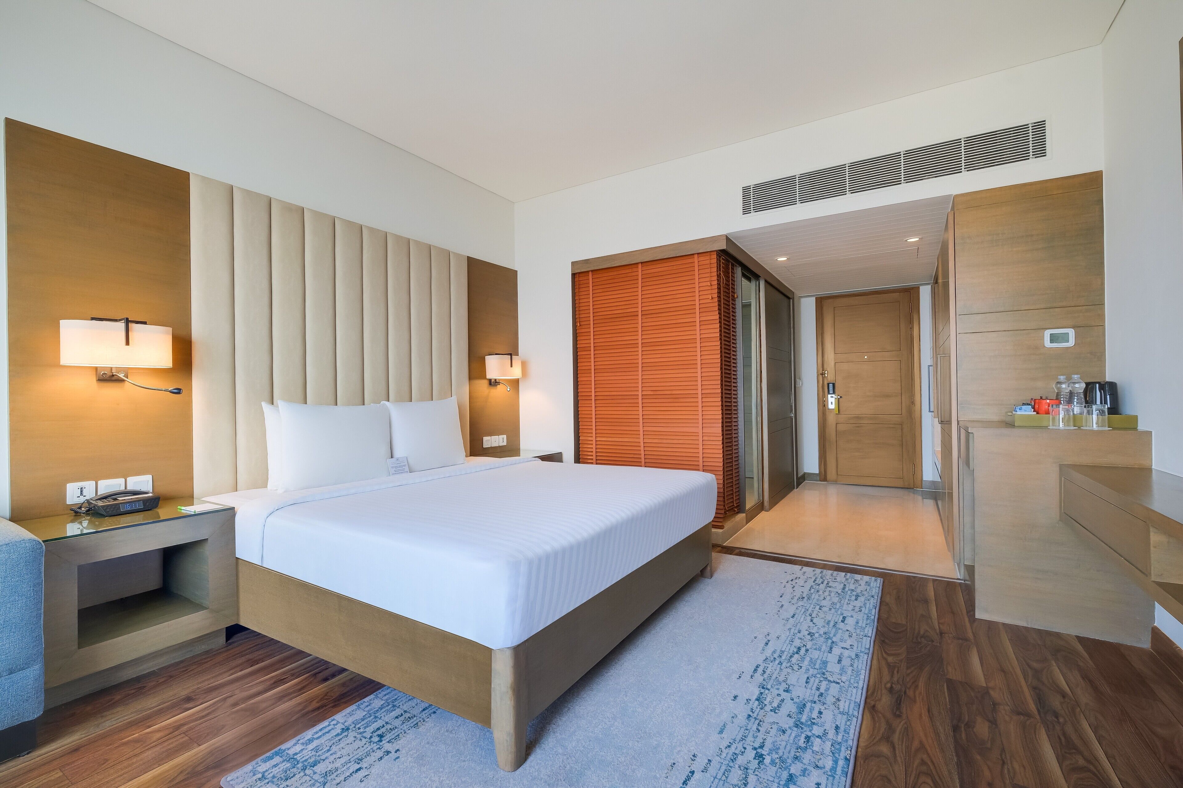 A modern hotel room with a large bed, bedside tables with lamps, a wooden headboard, a wardrobe, and a bathroom with a shower and toilet.