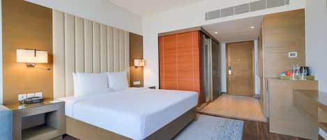 A modern hotel room with a large bed, bedside tables with lamps, a wooden headboard, a wardrobe, and a bathroom with a shower and toilet.
