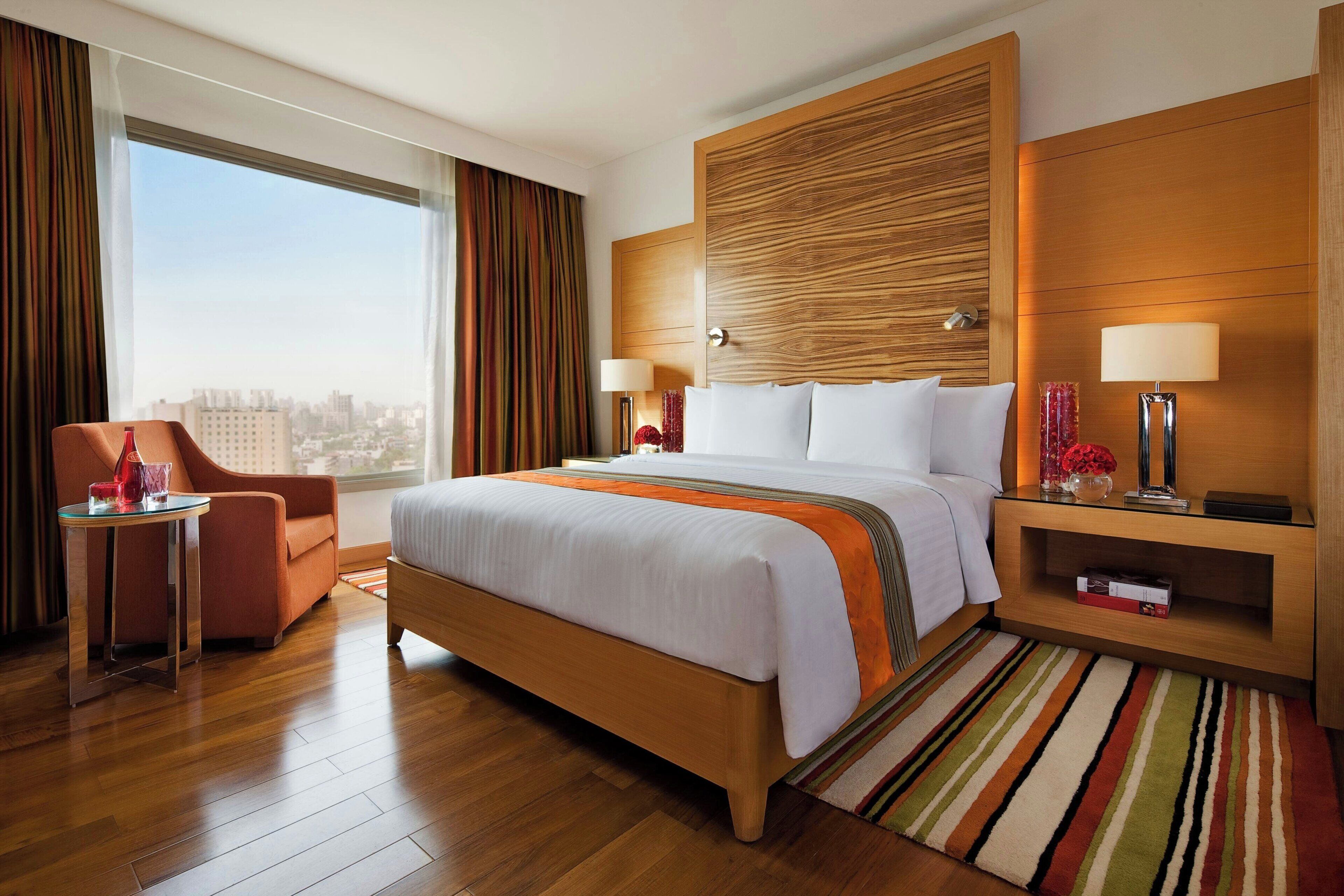 A hotel room with a large bed, a sitting area with a chair, a nightstand with a lamp, and a view of the city through the window.