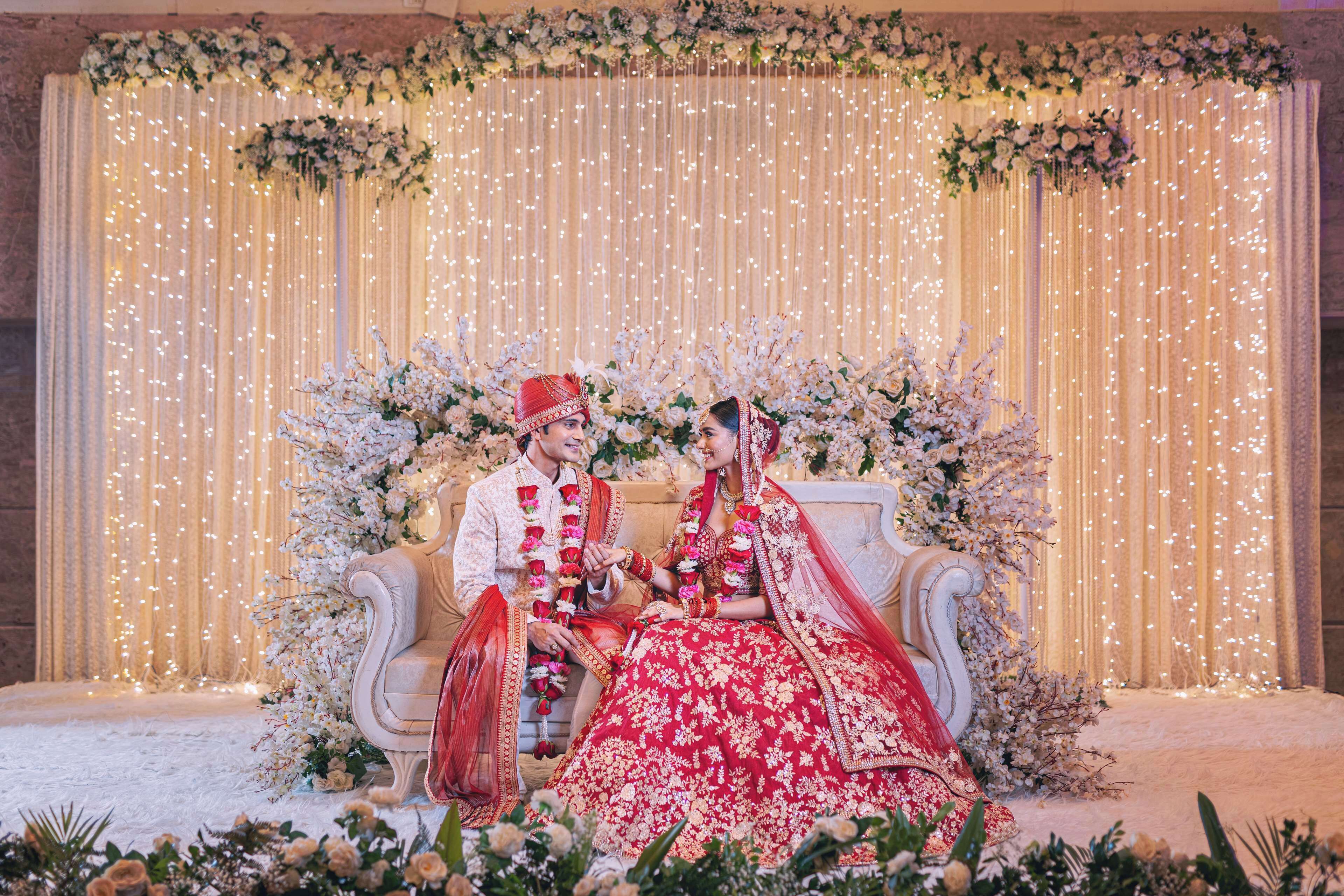 Una pareja sentada en un sofá ornamentado, con cortinas drapeadas y arreglos florales de fondo.