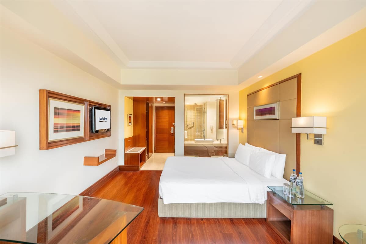 Een hotelkamer met een groot bed, een glazen tafel, een houten bureau, een spiegel en een lijst met een afbeelding aan de muur.