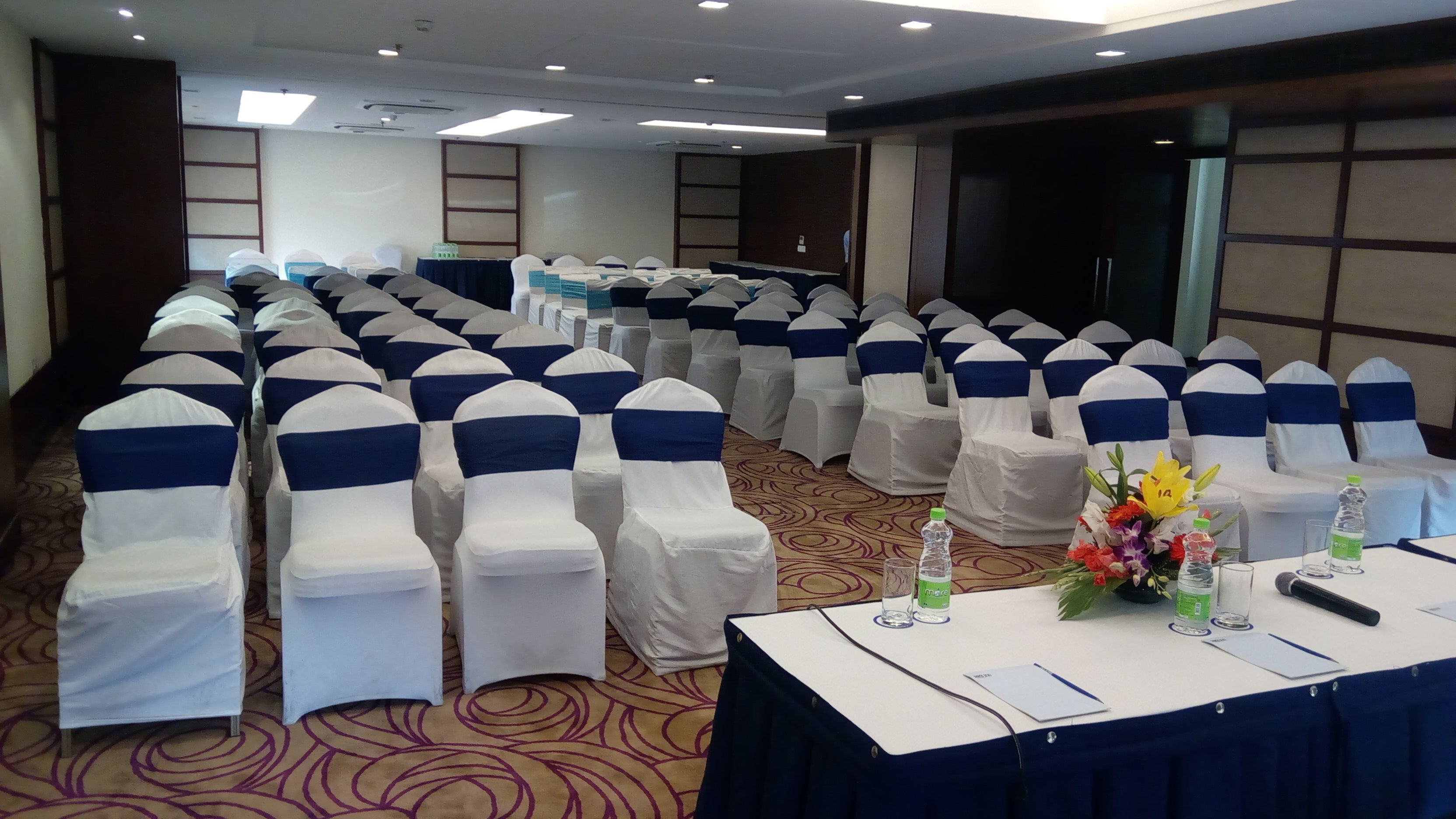 A conference room with rows of chairs covered in white and blue sashes, a table with water bottles and a flower arrangement, and a carpeted floor.