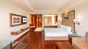 A modern hotel room with a large bed, a glass table, a sofa, wooden flooring, and a bathroom visible through an open door. - Park Inn, Gurgaon (Gurugram)