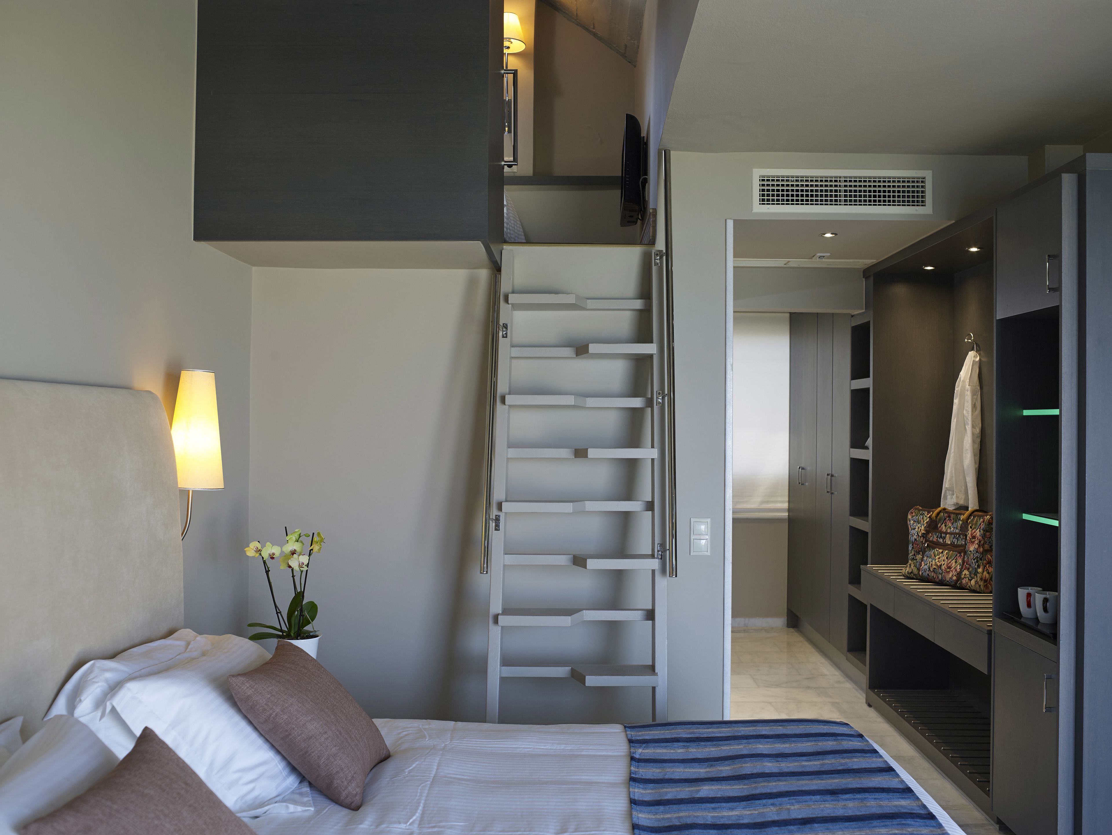 A modern hotel room with a bed, a bedside lamp, a potted plant, a built-in wardrobe, and a small seating area with a bench.