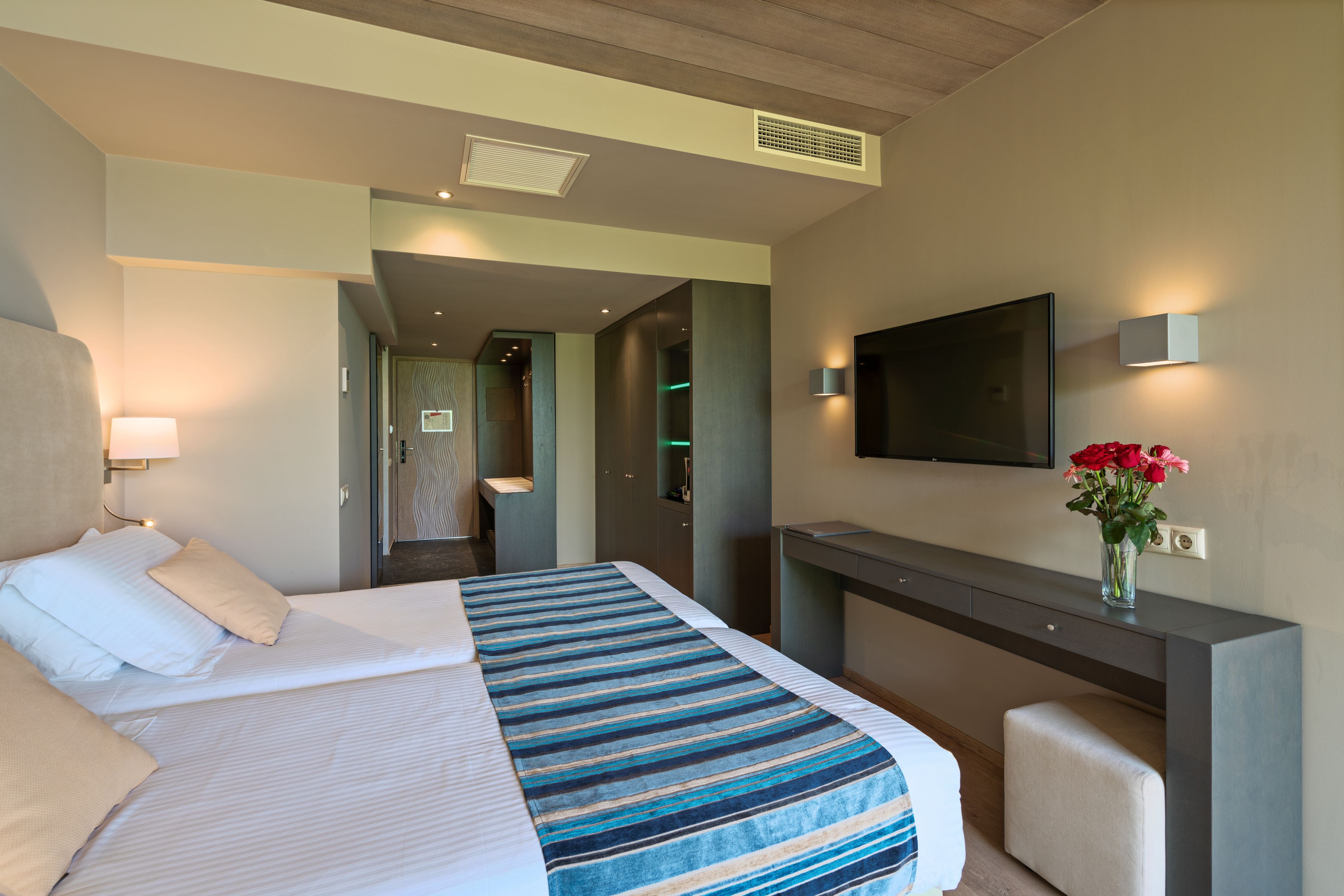 Una habitación de hotel con cama, escritorio, televisor y un florero con flores.