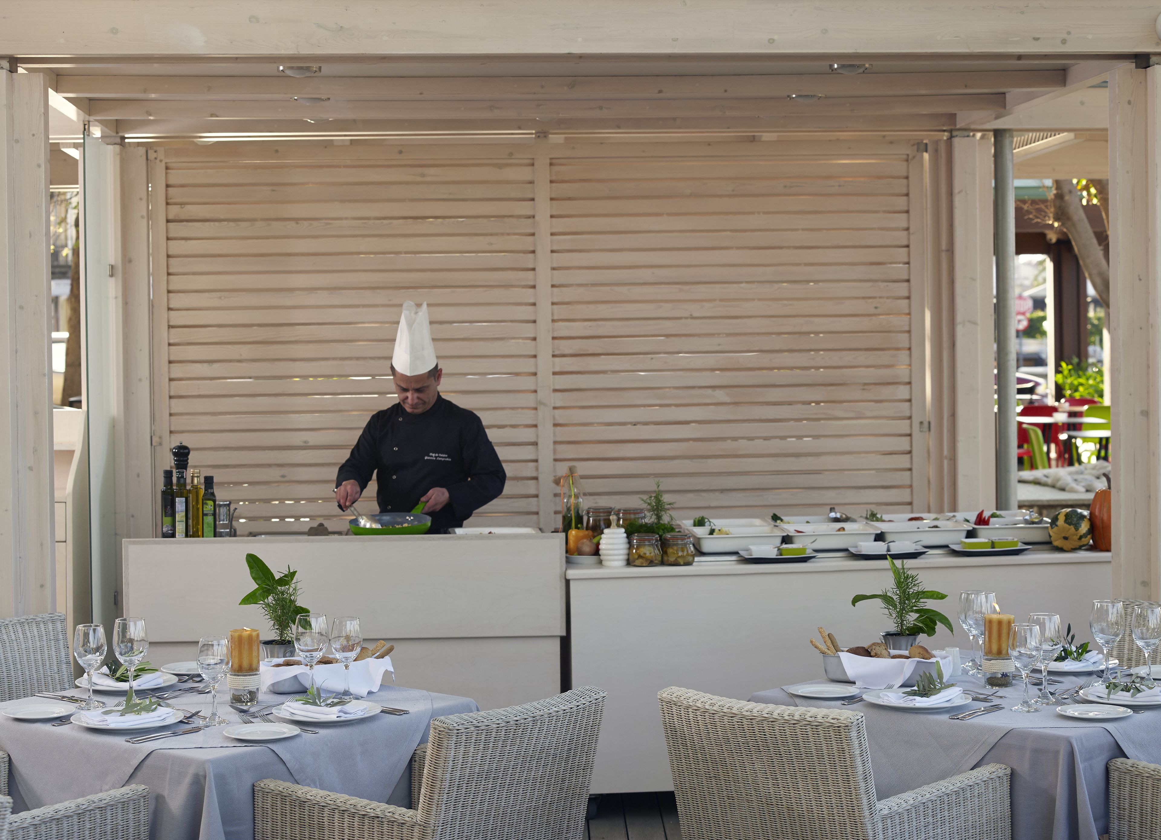 Un chef preparando comida en un comedor al aire libre con sillas y mesas de mimbre dispuestas para una comida.