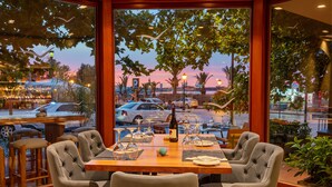 A restaurant with a large window view of a parking lot and a sunset. - Kyma Suites Beach Hotel (Rethymno)