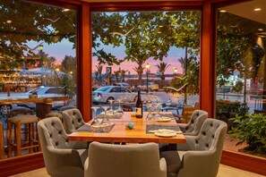 A restaurant with a large window view of a parking lot and a sunset. - Kyma Suites Beach Hotel (Rethymno)