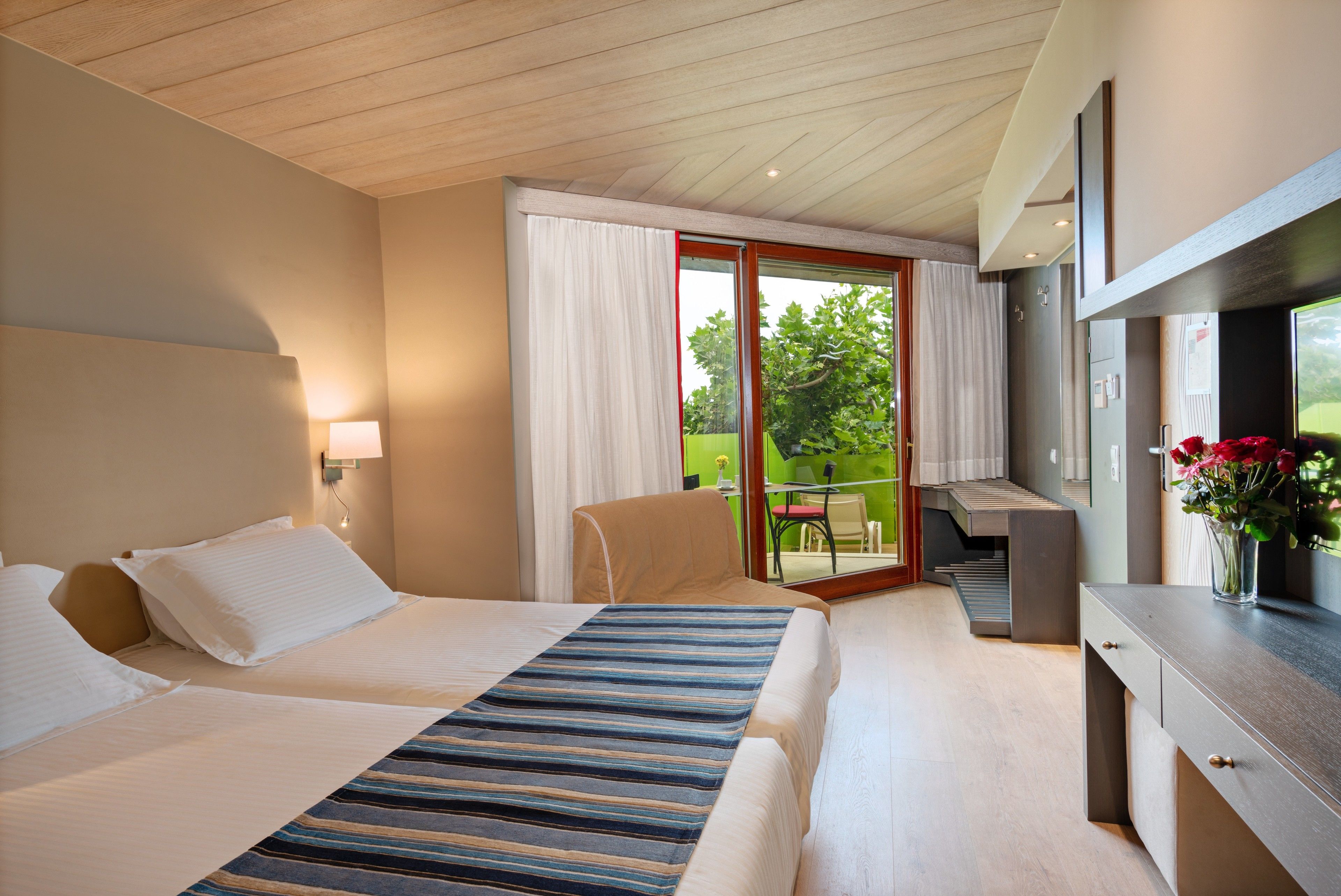 A hotel room with two beds, a desk, and a balcony with a view of greenery.