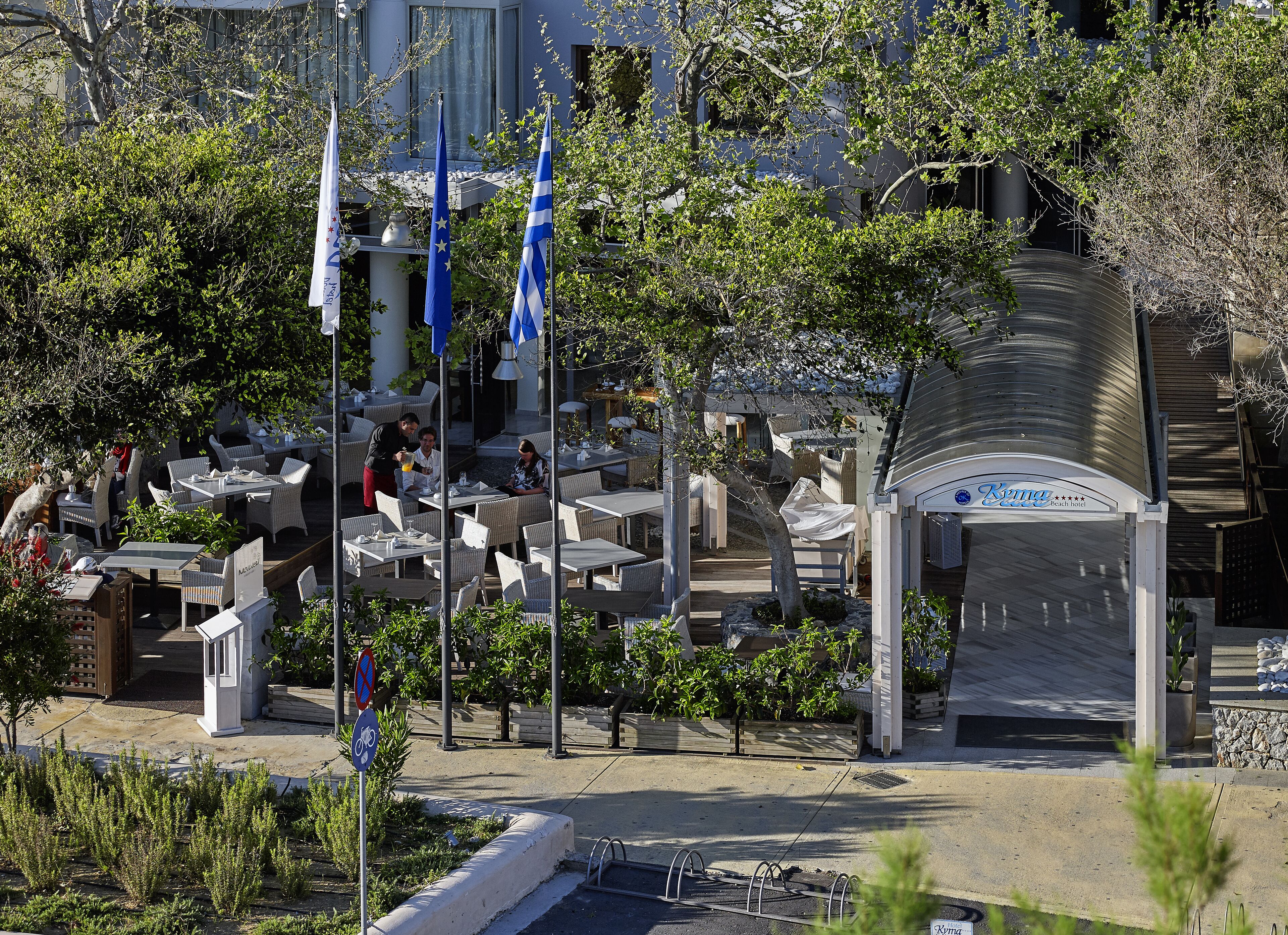 Un área de estar al aire libre con mesas y sillas, banderas y un edificio con un letrero que dice "Kymma beach hotel".