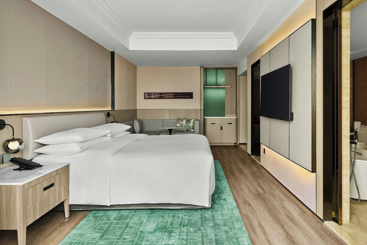 A modern hotel room with a large bed, a desk, a flat-screen TV, and a green patterned rug.