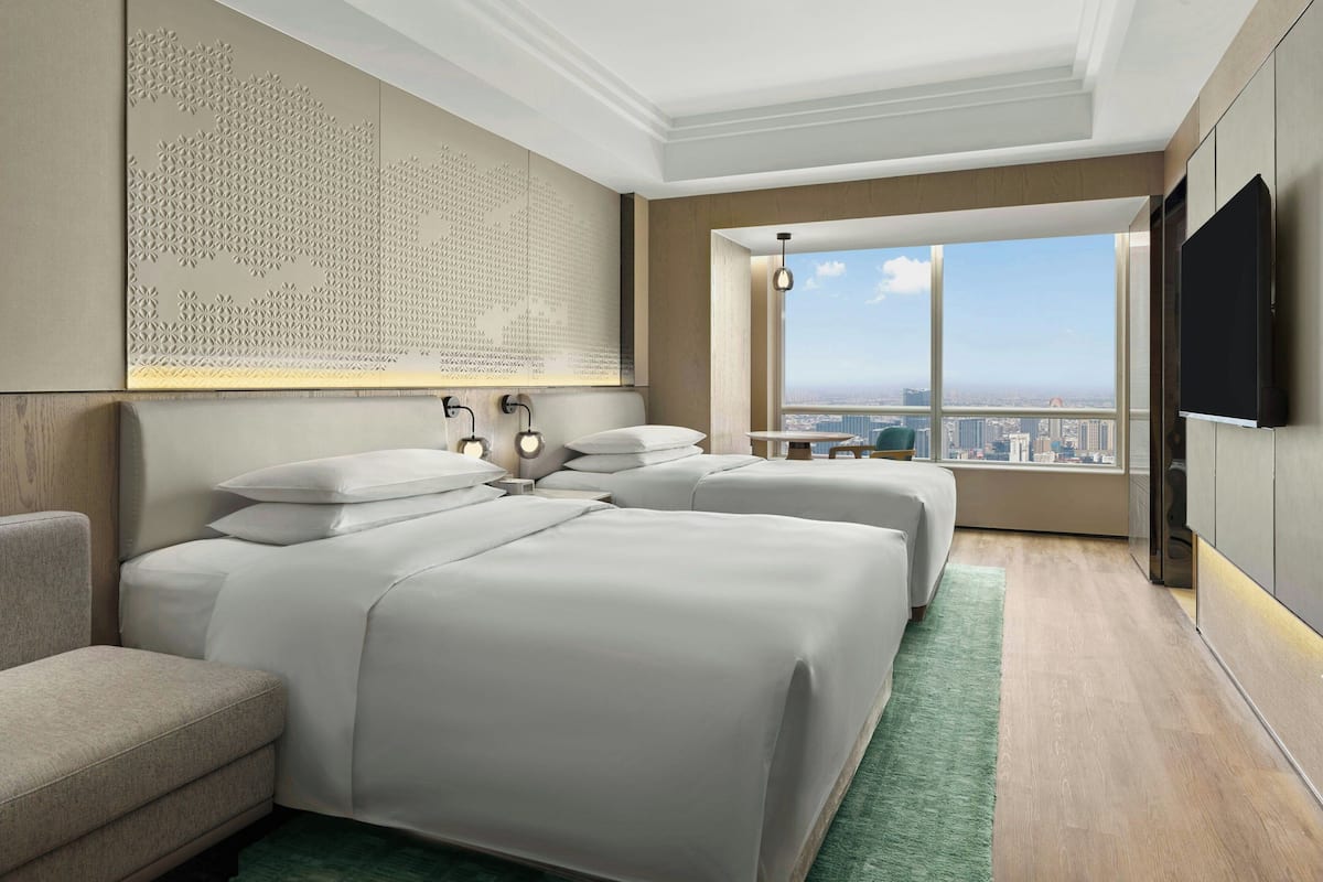 A modern hotel room with two beds, a sofa, a large window with a city view, and a mounted TV.
