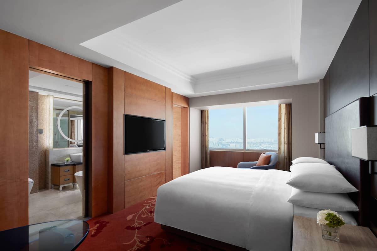 A hotel room with a large bed, a flat-screen TV, a desk with a chair, a view of the city, and a red carpet.