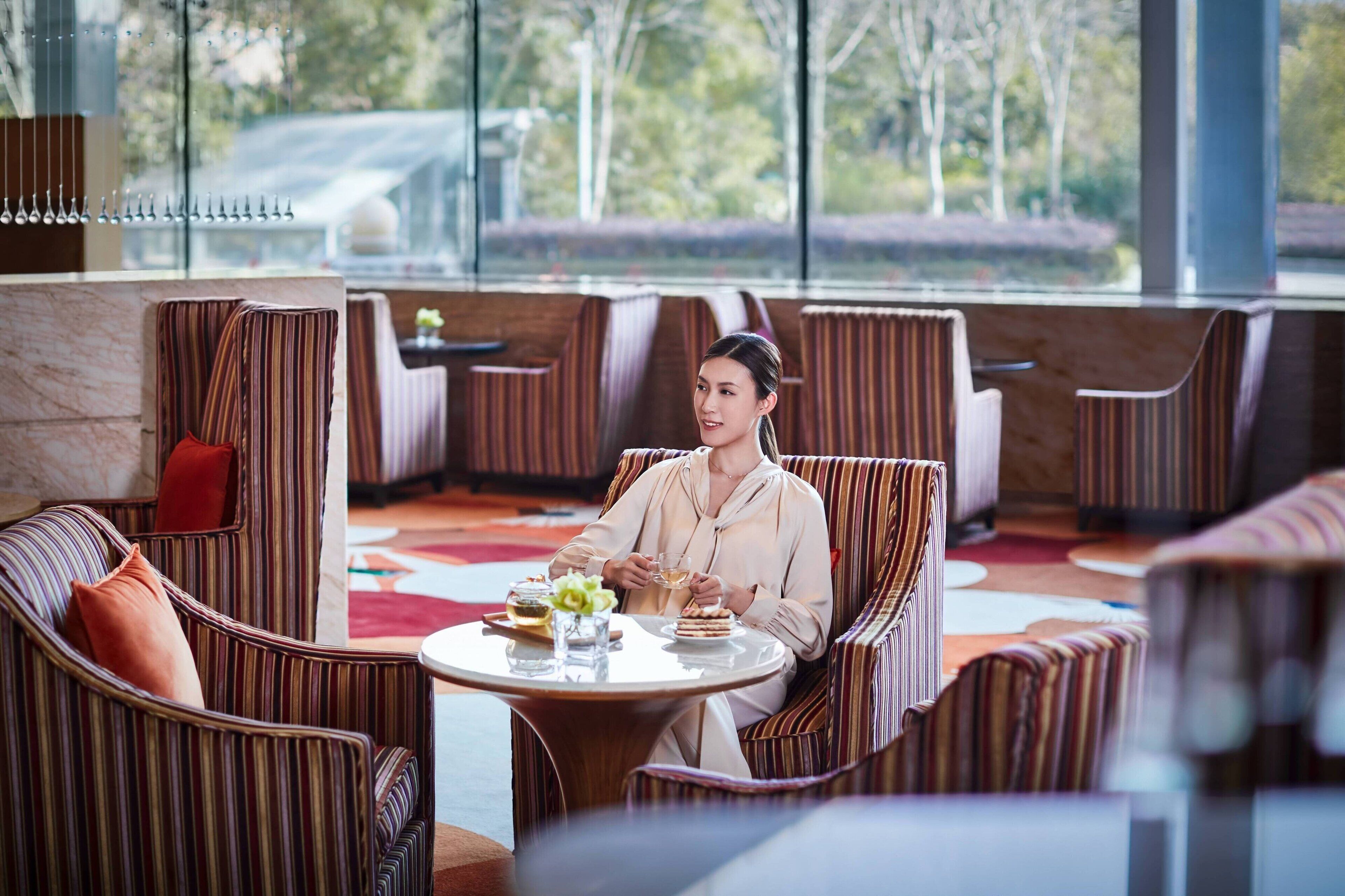A woman seated at a round table in a modern lounge area with striped upholstered chairs and a red cushion.