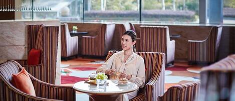 A woman seated at a round table in a modern lounge area with striped upholstered chairs and a red cushion.