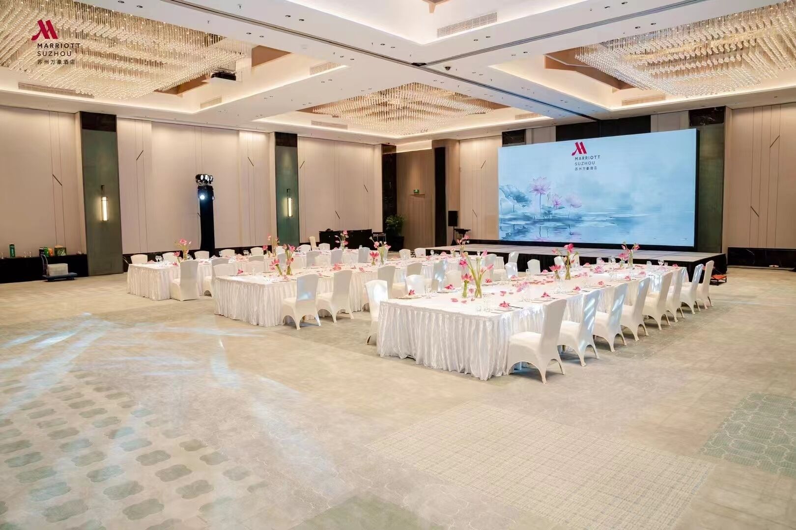 A large, well-lit conference room with multiple round tables, each set with white tablecloths and pink flowers.