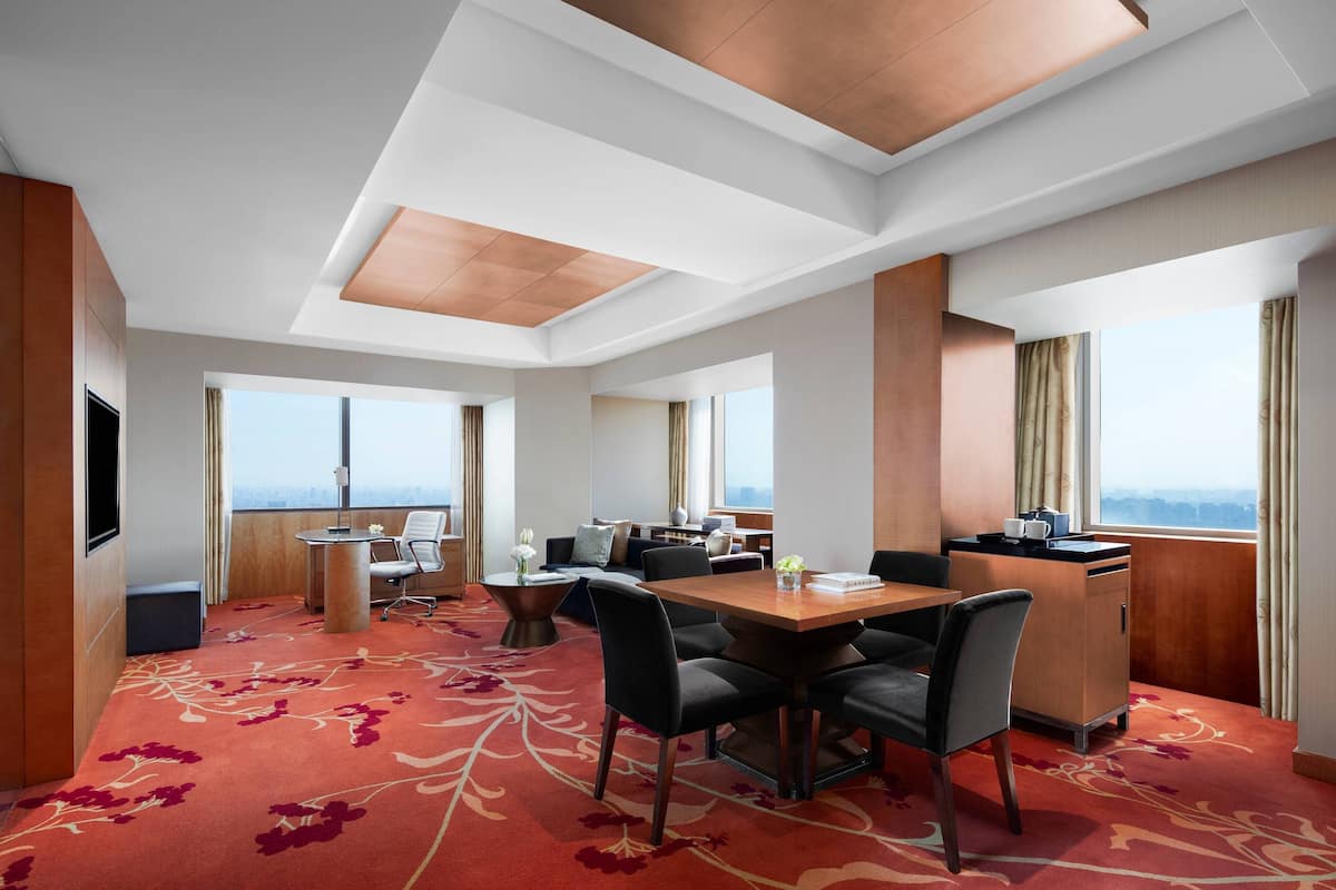 A hotel room with a dining area, a sofa, a TV, and large windows offering a city view.