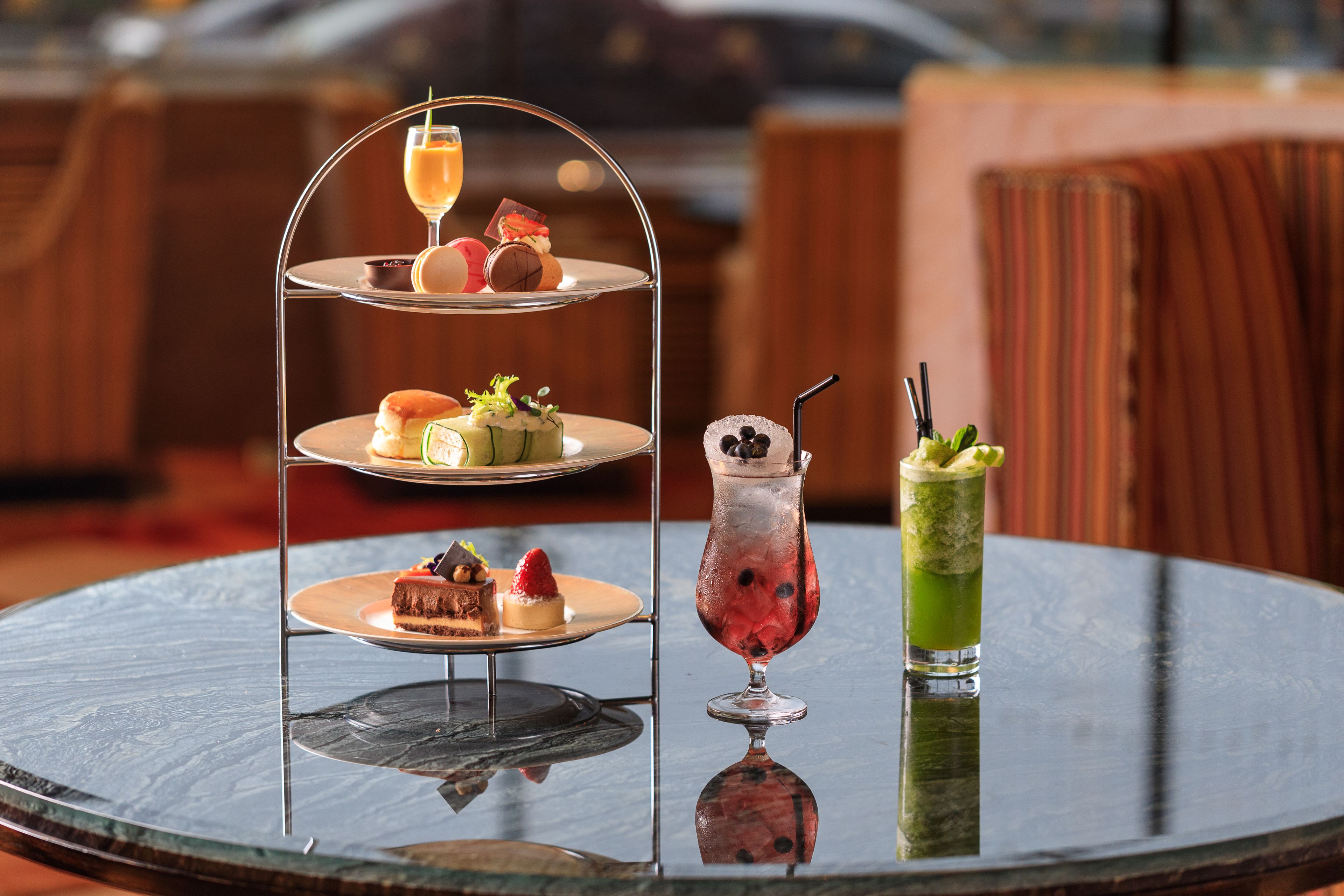 A three-tiered dessert stand with various pastries and a glass of a green beverage on a round table.