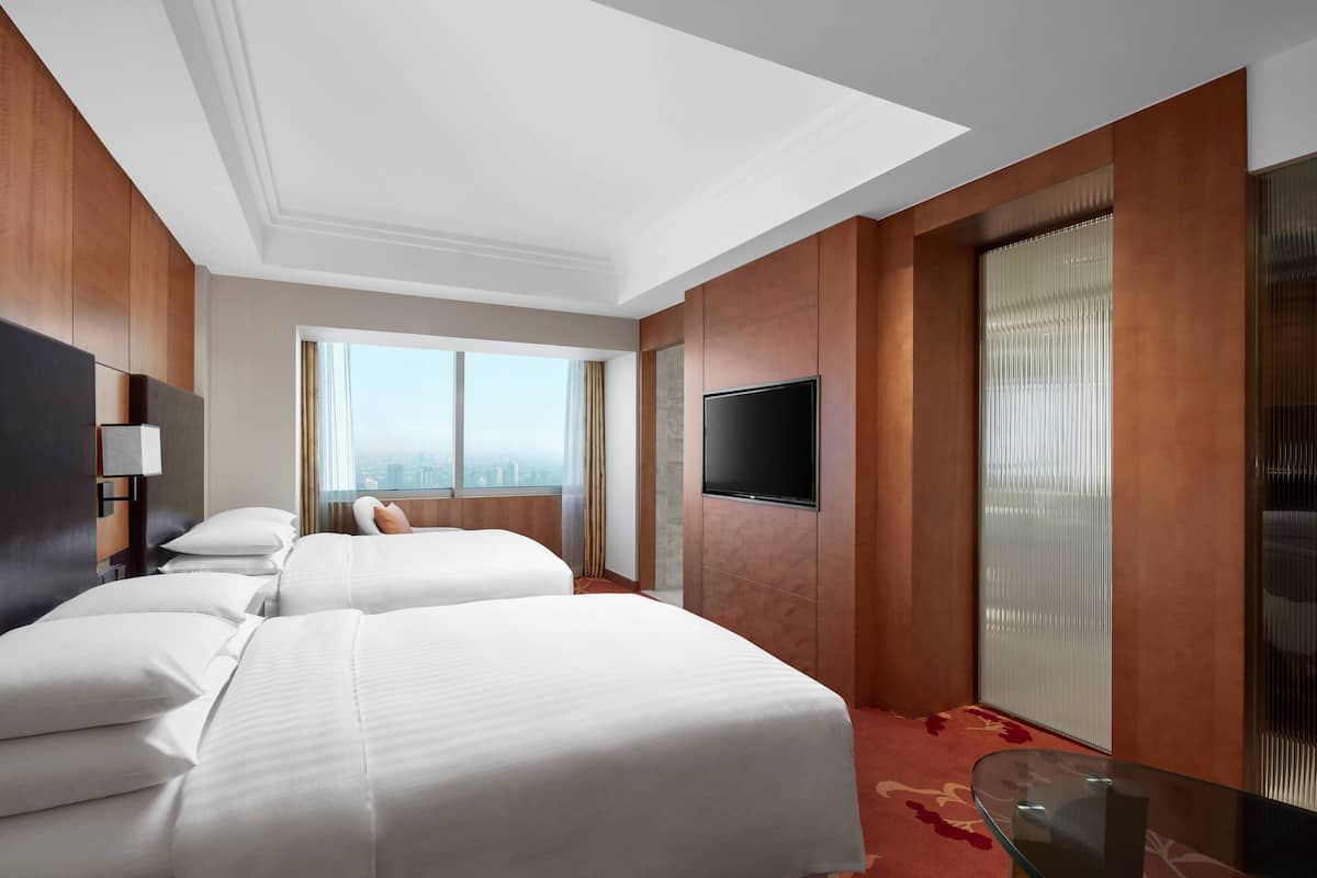A hotel room with two beds, a flat-screen TV, a glass table, and a city view through large windows.